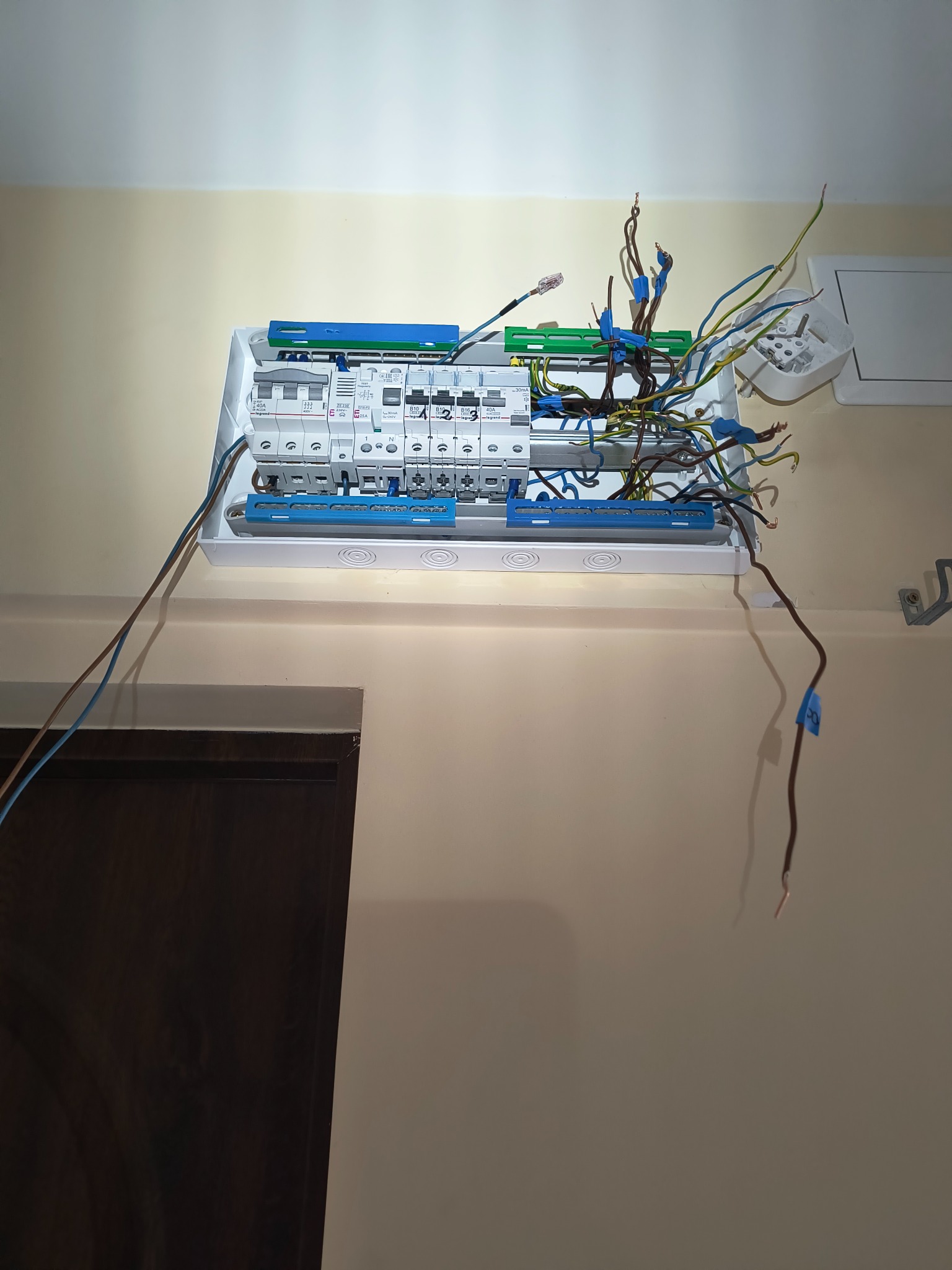 Otwarta skrzynka bezpiecznikowa z wystającymi przewodami w kolorach: zielonym, niebieskim i brązowym, zamontowana na ścianie wewnątrz budynku.