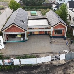 ERKABE SPÓŁKA Z OGRANICZONĄ ODPOWIEDZIALNOŚCIĄ - Budowa domu jednorodzinnego w stanie surowym otwartym, widok z lotu ptaka. Czerwona cegła, szara dachówka, widoczne materiały budowlane i reklama firmy.
