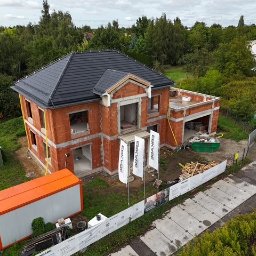 ERKABE SPÓŁKA Z OGRANICZONĄ ODPOWIEDZIALNOŚCIĄ - Dwupiętrowy dom w stanie surowym otwartym z czarnym dachem, widok z góry. Na działce kontener budowlany i materiały. W tle zieleń i zabudowania.