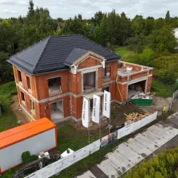 Dwupiętrowy dom w stanie surowym otwartym z czarnym dachem, widok z góry. Na działce kontener budowlany i materiały. W tle zieleń i zabudowania.