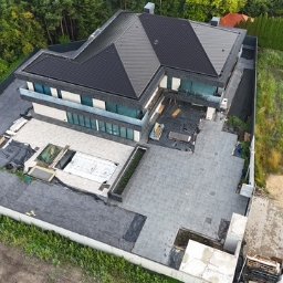 ERKABE SPÓŁKA Z OGRANICZONĄ ODPOWIEDZIALNOŚCIĄ - Nowoczesny dom z płaskim dachem, widok z góry. Częściowo wykończony taras z basenem. Widoczne samochody i zieleń w tle. Ujęcie z drona.