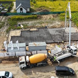 ERKABE SPÓŁKA Z OGRANICZONĄ ODPOWIEDZIALNOŚCIĄ - Wysoki kąt: wylewanie betonu na zbrojone fundamenty domu jednorodzinnego. Na pierwszym planie betonowóz z pompą. W tle dwa domy i łąka.