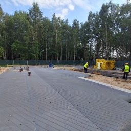 ERKABE SPÓŁKA Z OGRANICZONĄ ODPOWIEDZIALNOŚCIĄ - Przygotowanie terenu pod fundamenty: rozłożona geowłóknina, widoczne rury kanalizacyjne, pracownicy w odblaskowych kamizelkach. W tle las.
