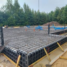ERKABE SPÓŁKA Z OGRANICZONĄ ODPOWIEDZIALNOŚCIĄ - Zbrojenie fundamentu z szalunkiem systemowym i instalacją wodną. Widok na przygotowany teren budowy z drzewami w tle.