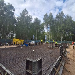 ERKABE SPÓŁKA Z OGRANICZONĄ ODPOWIEDZIALNOŚCIĄ - Zbrojenie płyty fundamentowej z szalunkiem metalowym, widoczni pracownicy w odblaskowych kamizelkach i żółtych kaskach. W tle drzewa i błękitne niebo.