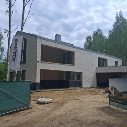 ERKABE SPÓŁKA Z OGRANICZONĄ ODPOWIEDZIALNOŚCIĄ - Nowoczesny budynek mieszkalny z elewacją z jasnej cegły klinkierowej w zaawansowanym etapie budowy we Wrocławiu, z dużymi oknami, płaskim dachem i otoczeniem zieleni. Widoczne ogrodzenie i flaga...