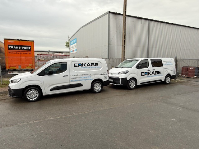 Dwa białe samochody dostawcze firmy budowlanej ERKABE z logo i danymi kontaktowymi, zaparkowane przed halą magazynową. Flota pojazdów budowlanych.