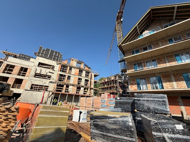 Budowa wielopiętrowego budynku z cegły, widoczny żuraw i materiały budowlane. Praca wre na budowie, w tle błękitne niebo.