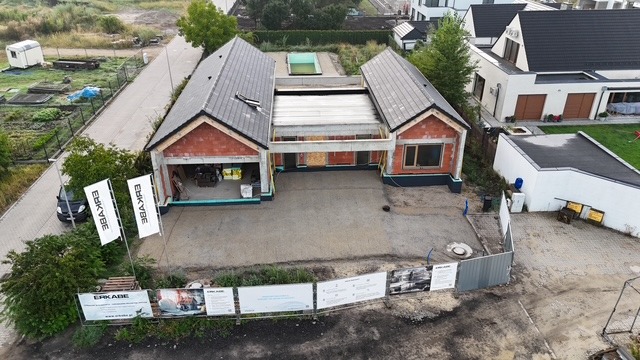 Budowa domu jednorodzinnego w stanie surowym otwartym, widok z lotu ptaka. Czerwona cegła, szara dachówka, widoczne materiały budowlane i reklama firmy.