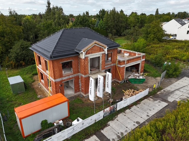 Dwupiętrowy dom w stanie surowym otwartym z czarnym dachem, widok z góry. Na działce kontener budowlany i materiały. W tle zieleń i zabudowania.