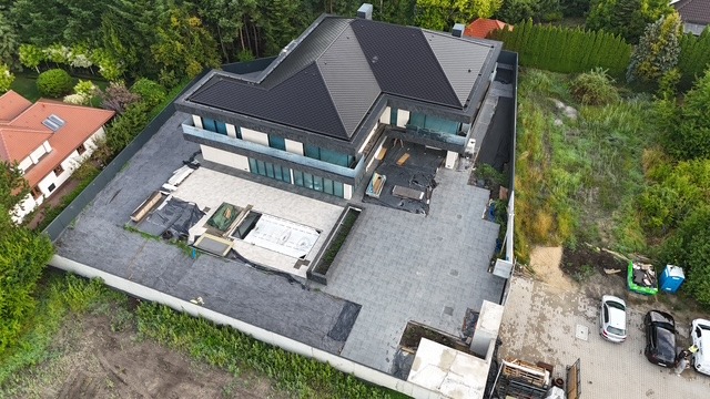 Nowoczesny dom z płaskim dachem, widok z góry. Częściowo wykończony taras z basenem. Widoczne samochody i zieleń w tle. Ujęcie z drona.