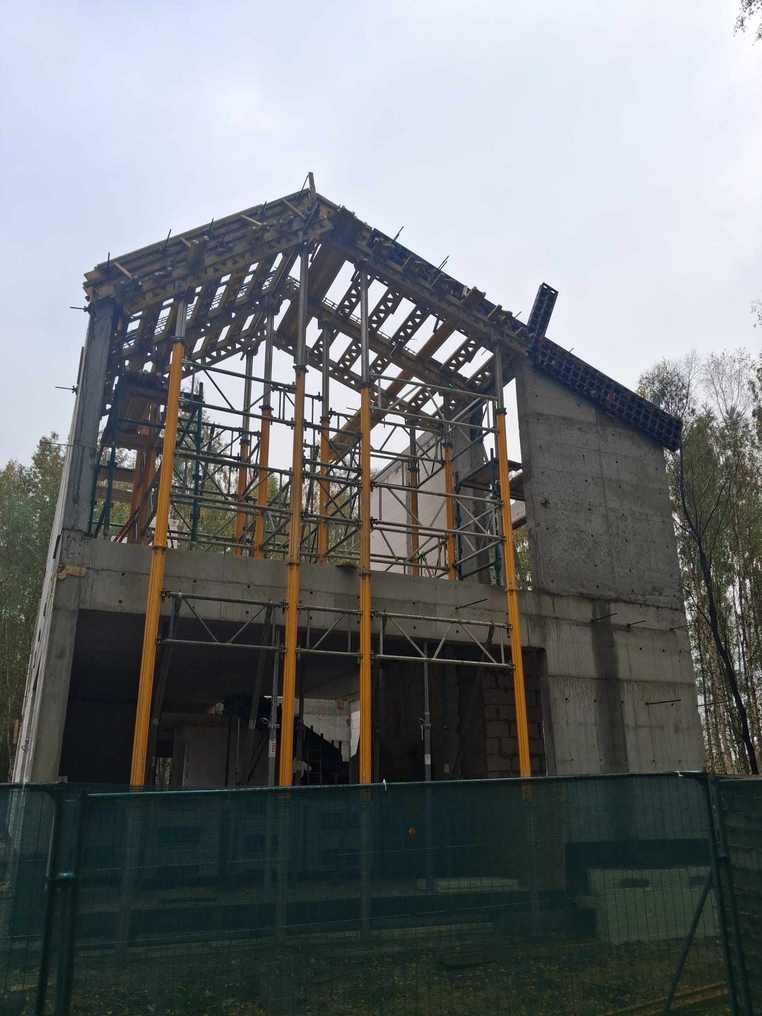 Budowa domu żelbetowego w stanie surowym otwartym: widoczne szalunki, zbrojenia i betonowe ściany. Ujęcie z dołu, z zielonym ogrodzeniem w tle i drzewami.