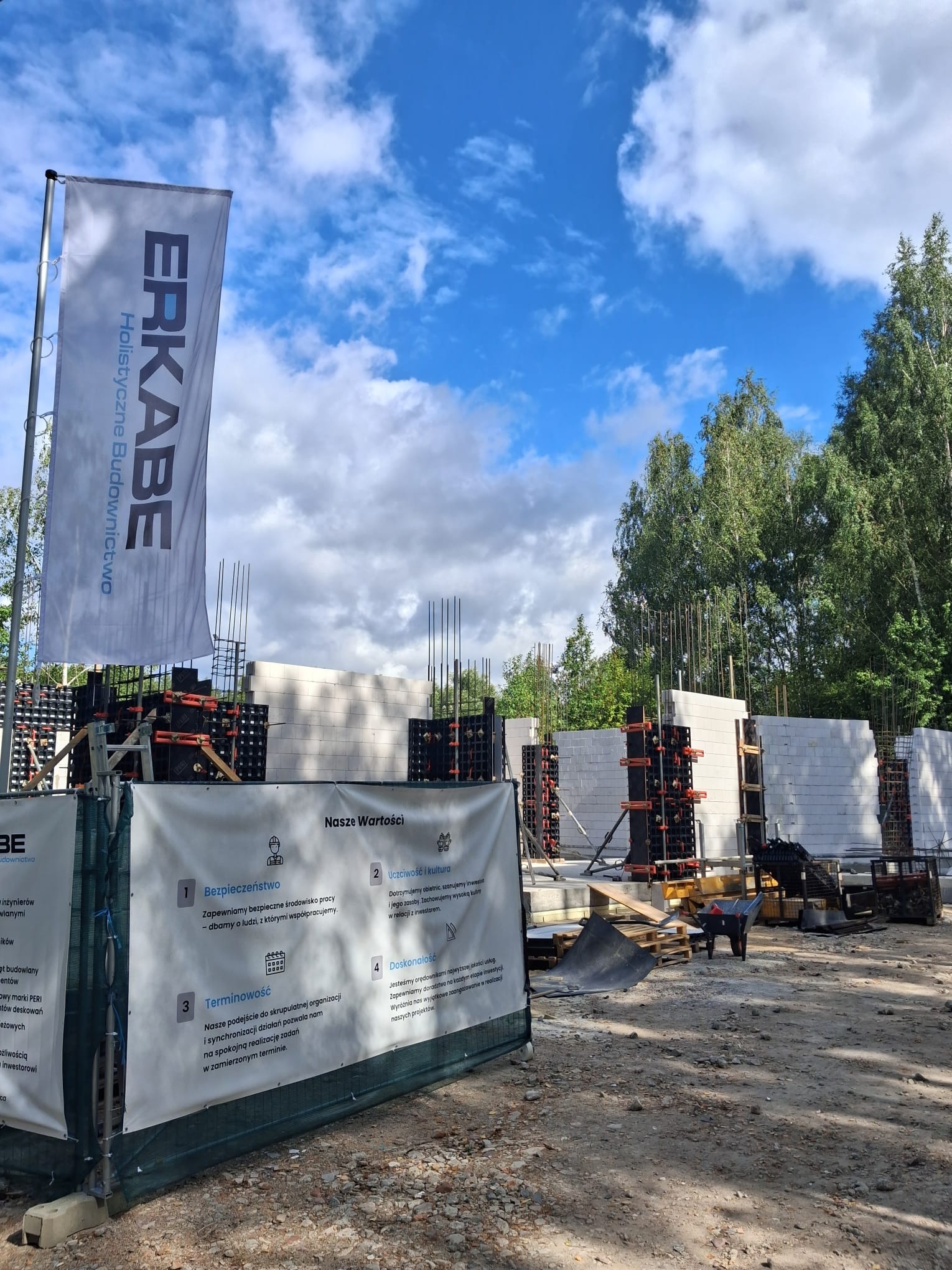 Budowa domu z betonu komórkowego w trakcie realizacji. Widoczne zbrojenia, szalunki i baner reklamowy firmy budowlanej. Błękitne niebo w tle.