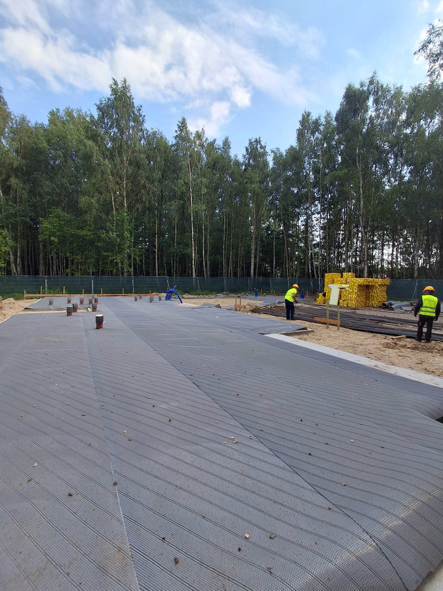 Przygotowanie terenu pod fundamenty: rozłożona geowłóknina, widoczne rury kanalizacyjne, pracownicy w odblaskowych kamizelkach. W tle las.