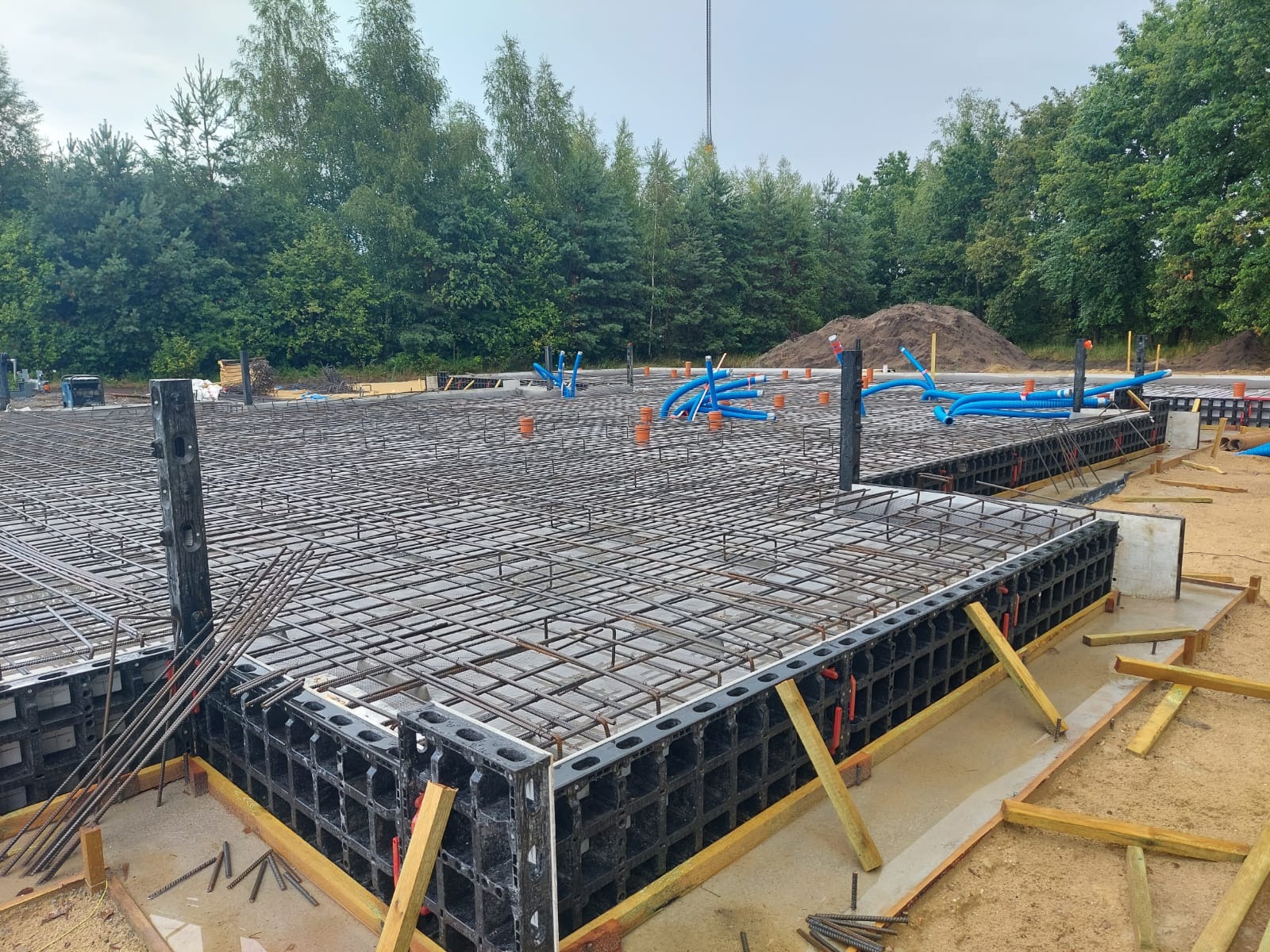 Zbrojenie fundamentu z szalunkiem systemowym i instalacją wodną. Widok na przygotowany teren budowy z drzewami w tle.