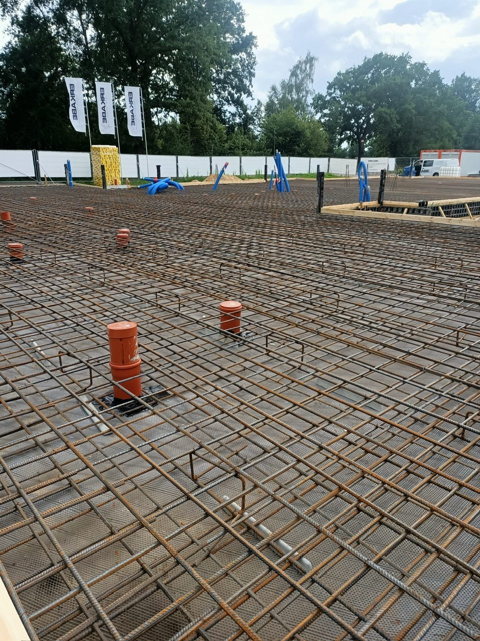 Zbrojenie fundamentu pod budowę domu jednorodzinnego z widocznymi elementami instalacji kanalizacyjnej i deskowaniem krawędzi. Na dalszym planie drzewa i ogrodzenie.