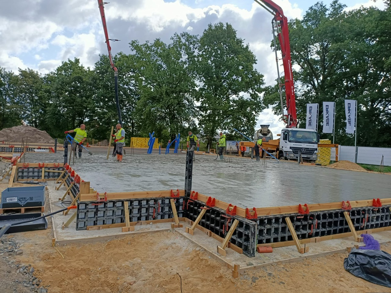 Wylewanie betonu na przygotowany fundament z szalunkami systemowymi. Widoczni pracownicy w żółtych kamizelkach i pompa do betonu. W tle drzewa i ciężarówka.
