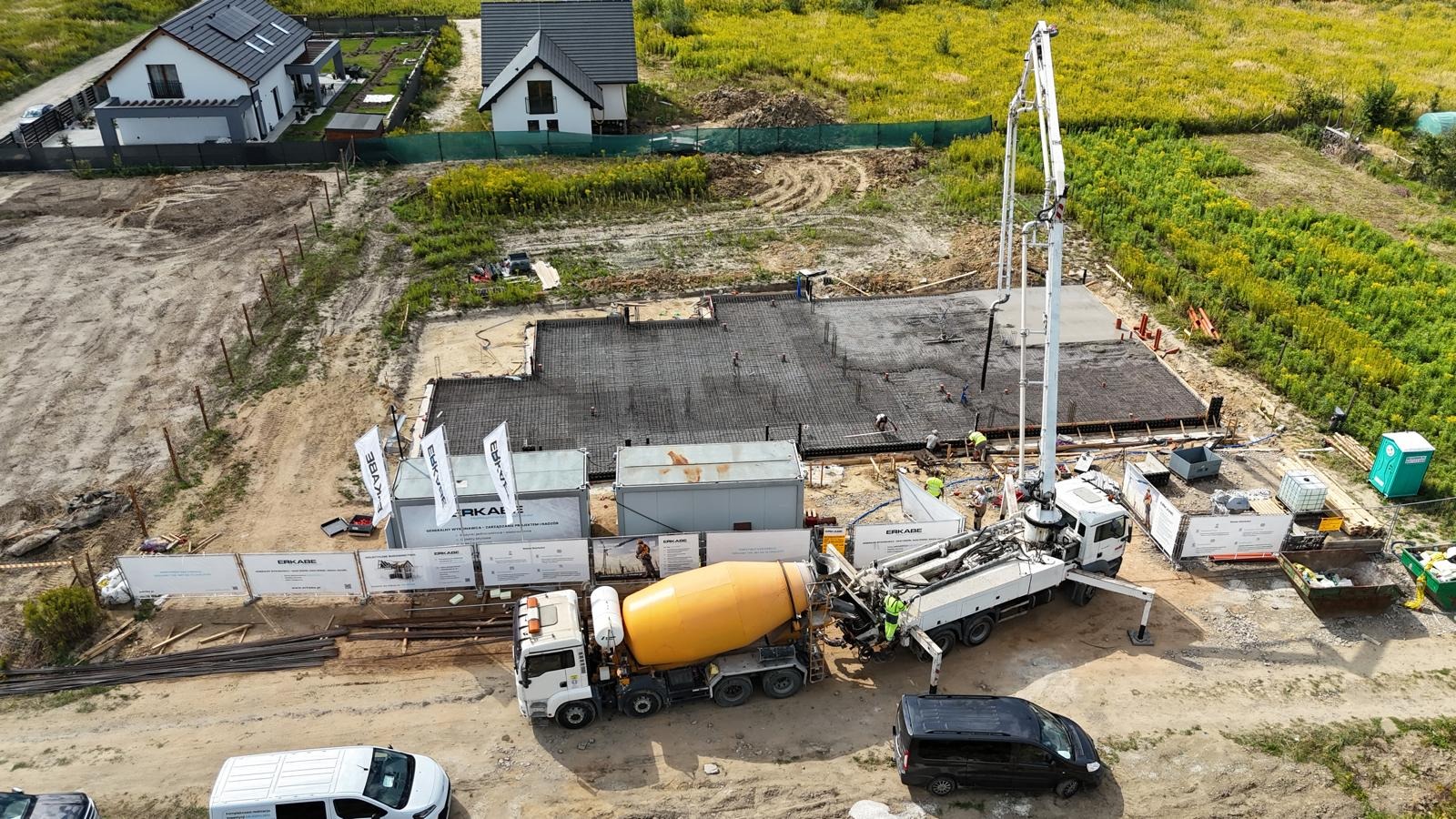 Wylewanie betonu na zbrojone fundamenty domu jednorodzinnego. Na budowie obecny betonowóz z pompą. Widok z góry na plac budowy z zielonym otoczeniem i zabudową.
