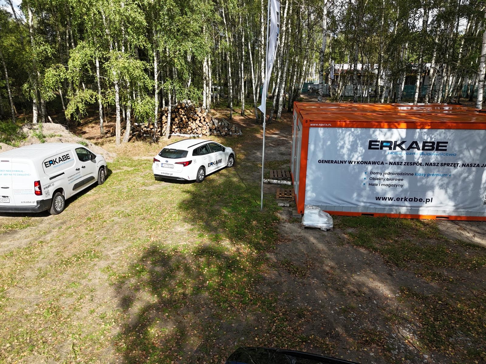 Plenerowe biuro budowy we Wrocławiu: Kontener biurowy, samochód osobowy i dostawczy z logo firmy budowlanej na tle lasu brzozowego i sterty drewna.