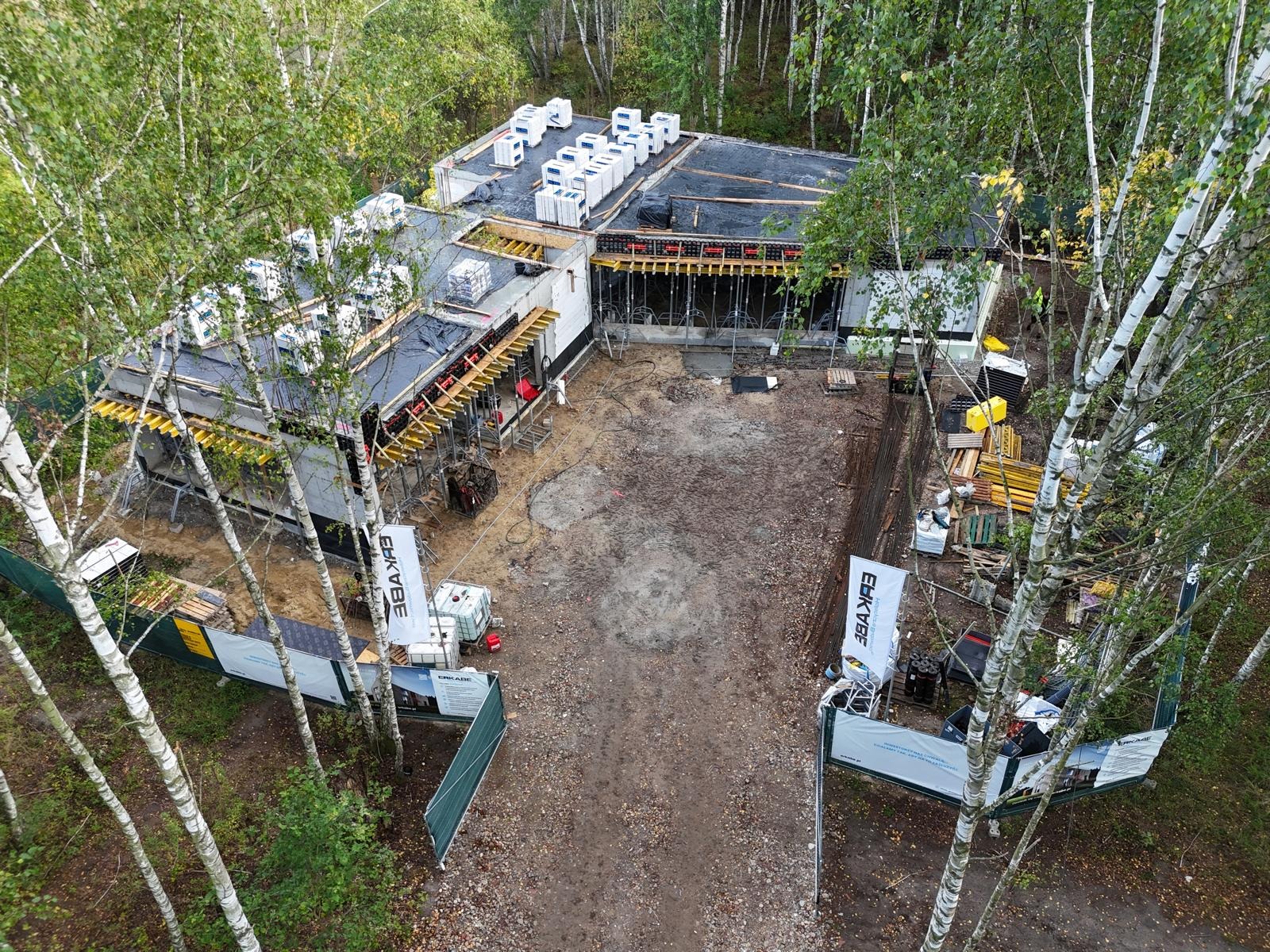 Budowa domu w lesie, widok z góry. Widać fundamenty, ściany w szalunkach, materiały budowlane i białe brzozy. Etap prac w toku, teren budowy zabezpieczony.