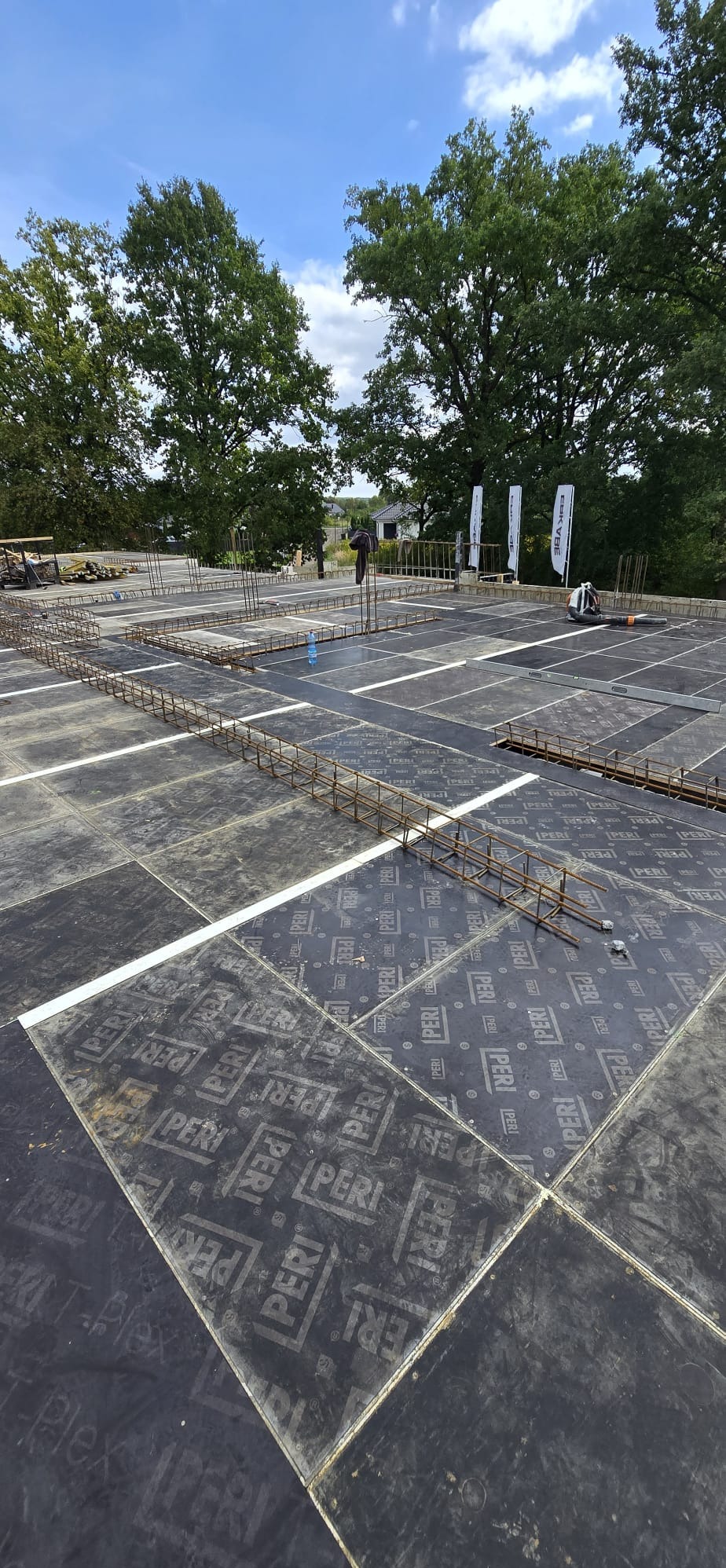 Zbrojenie fundamentów na czarnych płytach PERI, widok z góry. Konstrukcja z prętów stalowych przygotowana do zalania betonem. W tle drzewa i niebieskie niebo.