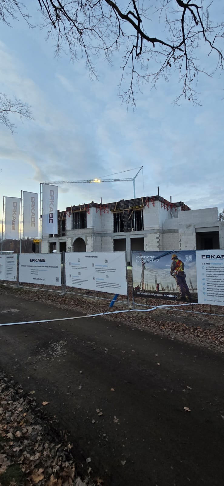 Budowa domu z betonu komórkowego w trakcie realizacji, widoczne szalunki i zbrojenia. Na pierwszym planie ogrodzenie z banerami reklamowymi firmy budowlanej.