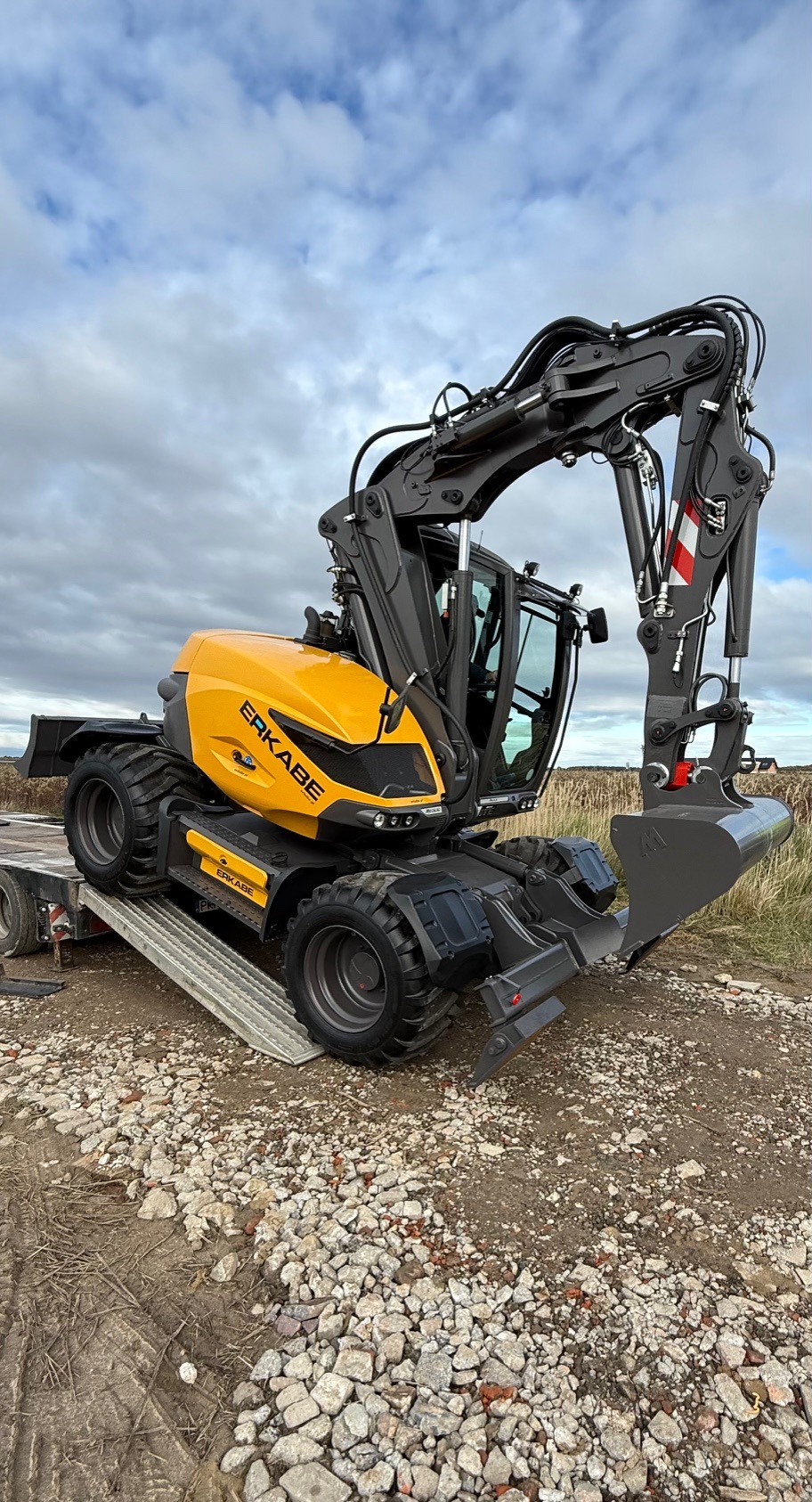 Żółto-szara koparka kołowa ER KABE na lawecie, widok z boku na tle pochmurnego nieba. Maszyna gotowa do transportu na plac budowy.
