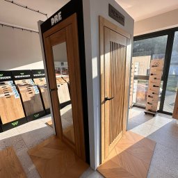 KINGA WŁODEK - Ekspozycja dwóch modeli drzwi wewnętrznych z drewna z czarnymi klamkami, na podłodze ułożonej w jodełkę. W tle widoczne wzorniki podłóg i ekspozytor klamek.