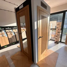 Ekspozycja dwóch modeli drzwi wewnętrznych z drewna z czarnymi klamkami, na podłodze ułożonej w jodełkę. W tle widoczne wzorniki podłóg i ekspozytor klamek.