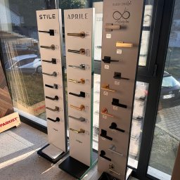 KINGA WŁODEK - Ekspozycja klamek drzwiowych marek Stile, Aprile i Infinity Line w salonie. Różne modele i wykończenia, od czarnych po złote, na tle okna i podłogi.
