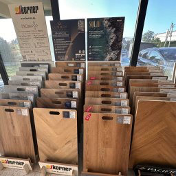 KINGA WŁODEK - Ekspozycja paneli podłogowych SPC w sklepie: różne wzory drewna, odcienie szarości i beżu. Panele z uchwytami, etykietami, na stojakach z logo Korner. Tło: okno, ulica.