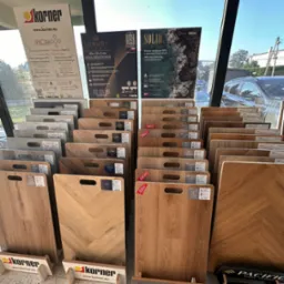 Ekspozycja paneli podłogowych SPC w sklepie: różne wzory drewna, odcienie szarości i beżu. Panele z uchwytami, etykietami, na stojakach z logo Korner. Tło: okno, ulica.