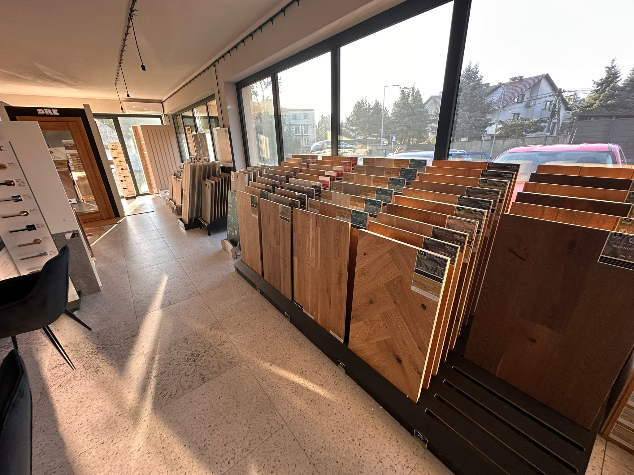 Salon z ekspozycją paneli podłogowych o różnorodnych wzorach i kolorach drewna, ustawionych pionowo na stojakach. Widoczne drzwi w tle.