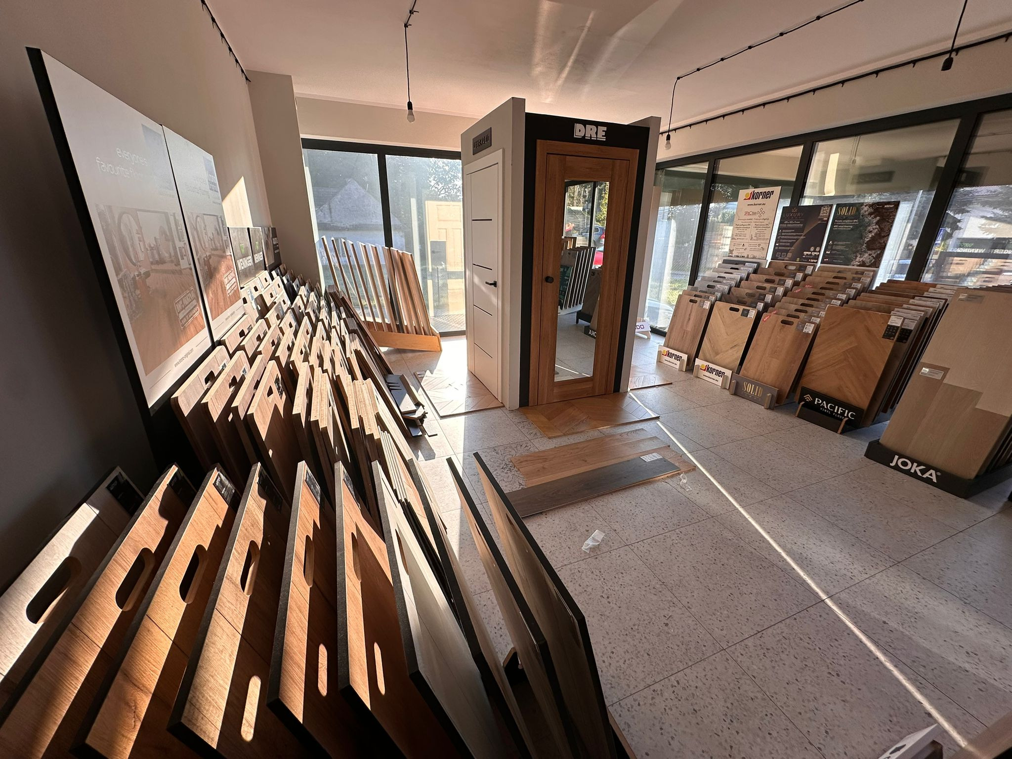 Ekspozycja w salonie: panele podłogowe Joka, Pacific, Korner oraz drzwi DRE. W tle widoczne próbki paneli ułożone przy oknie. Jasne wnętrze, duży wybór wzorów.