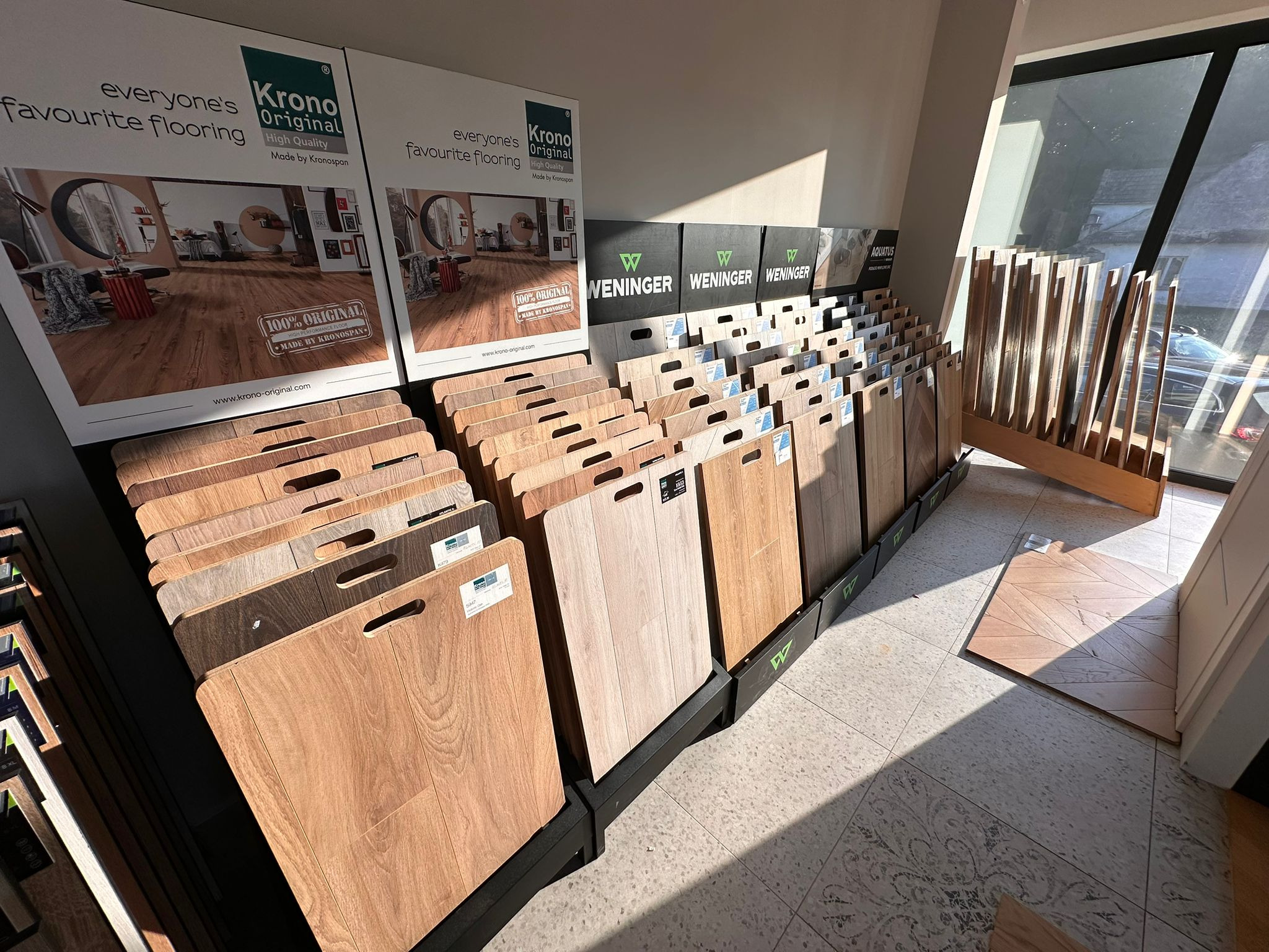Ekspozycja paneli podłogowych różnych producentów (Krono Original, Weninger, Aquatus) w salonie, z widocznymi próbkami i reklamami. Jasne światło dzienne.