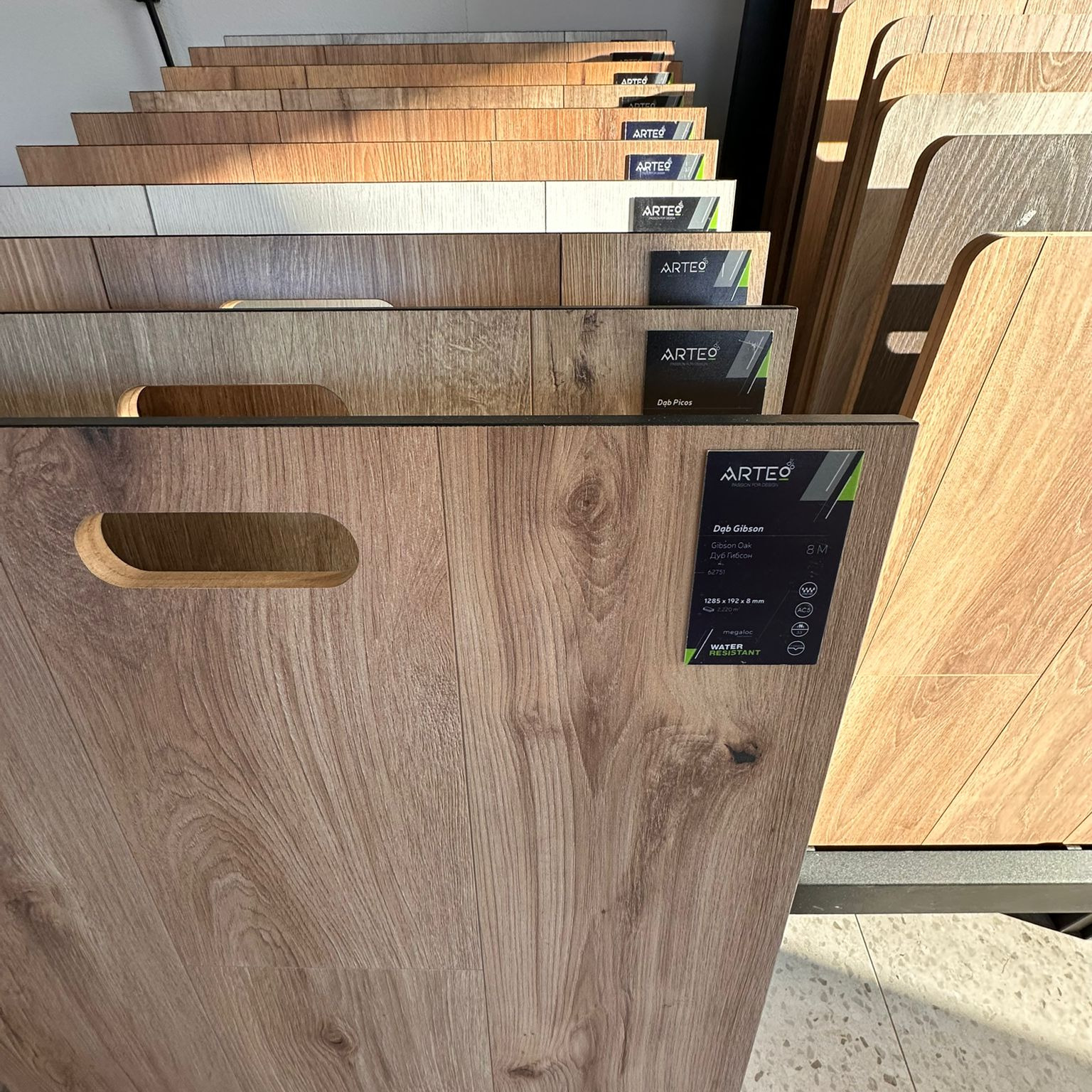 Ekspozycja paneli podłogowych Arteo w różnych odcieniach dębu, z widocznymi etykietami i specyfikacją techniczną, prezentowanych w salonie sprzedaży.