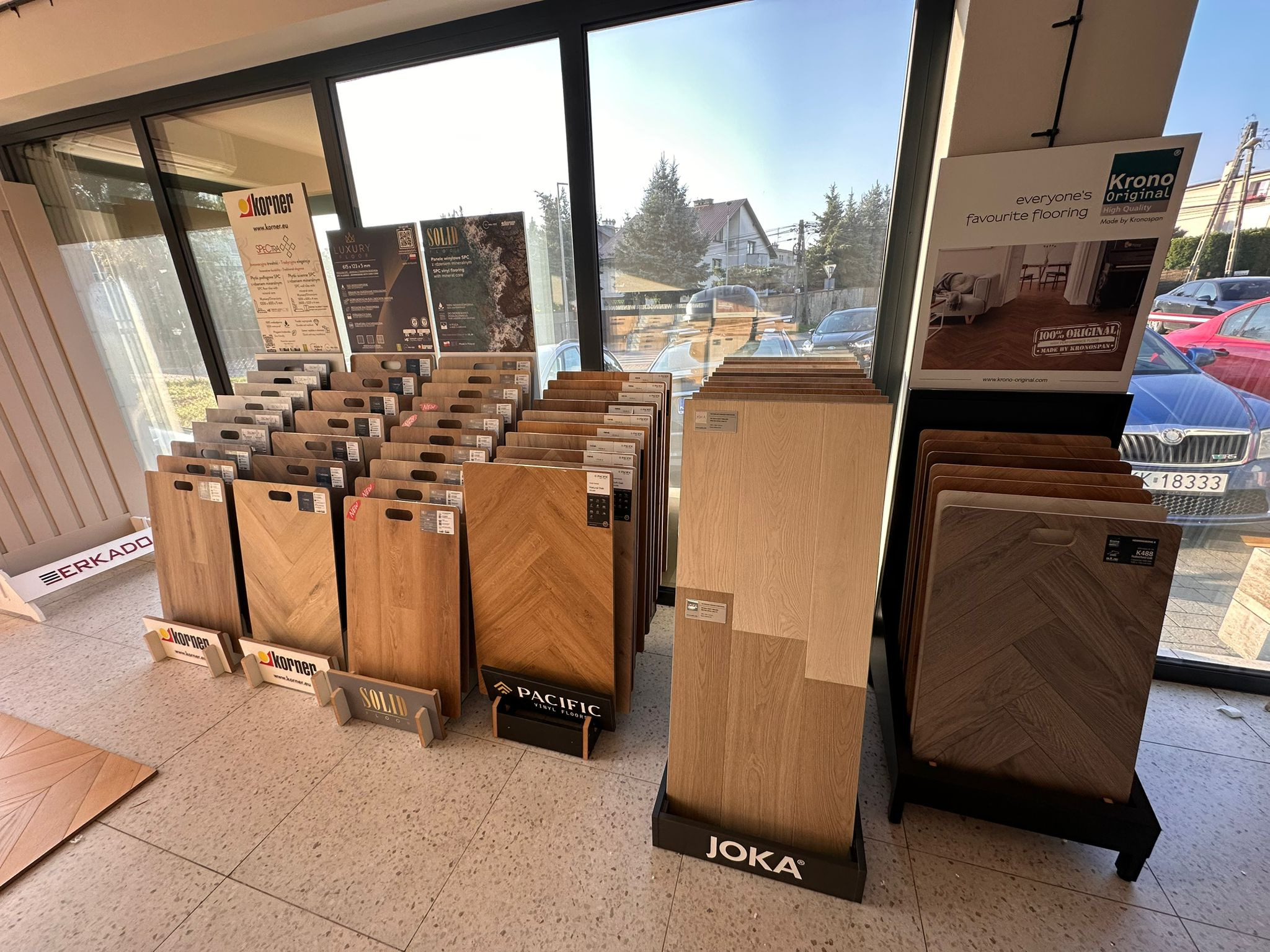 Ekspozycja paneli podłogowych różnych marek i wzorów w salonie sprzedaży, z widokiem na ulicę przez okno. Panele ułożone w stojakach, prezentujące różne odcienie drewna.