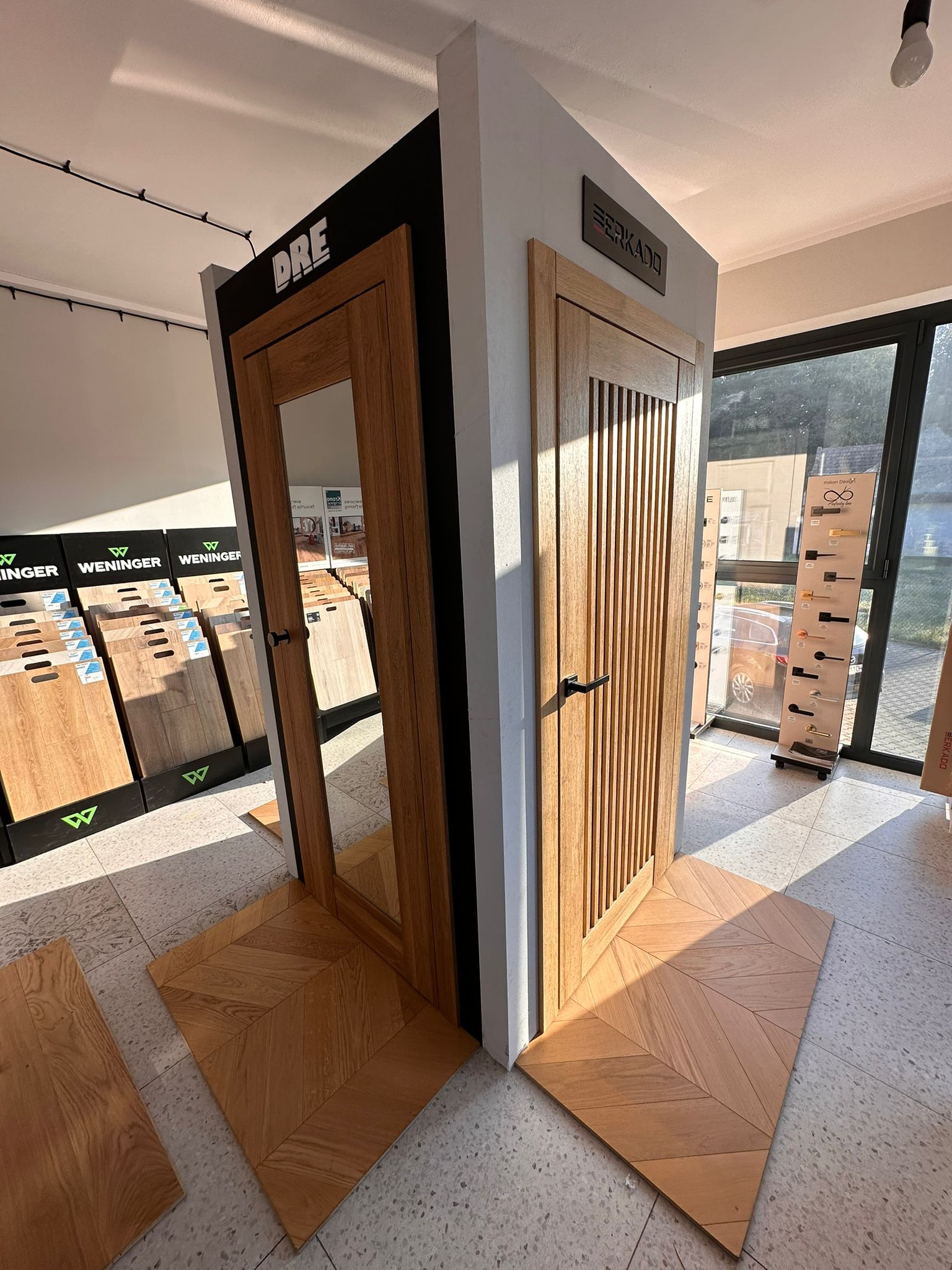 Ekspozycja dwóch modeli drzwi wewnętrznych z drewna z czarnymi klamkami, na podłodze ułożonej w jodełkę. W tle widoczne wzorniki podłóg i ekspozytor klamek.