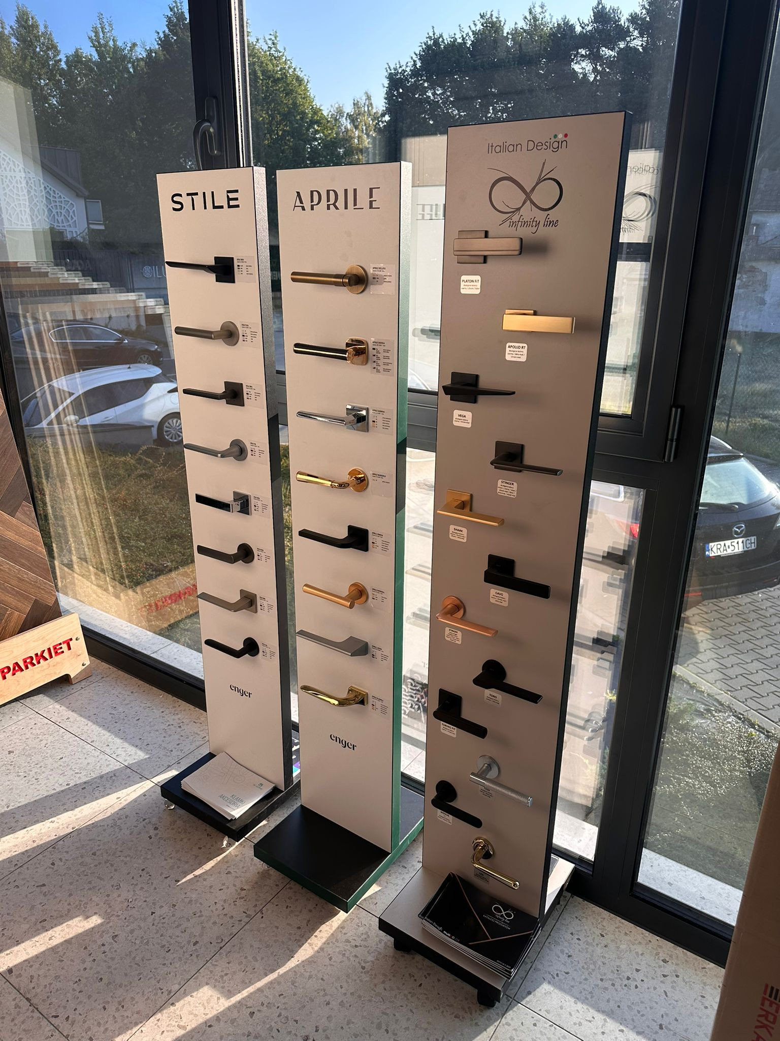 Ekspozycja klamek drzwiowych marek Stile, Aprile i Infinity Line w salonie. Różne modele i wykończenia, od czarnych po złote, na tle okna i podłogi.