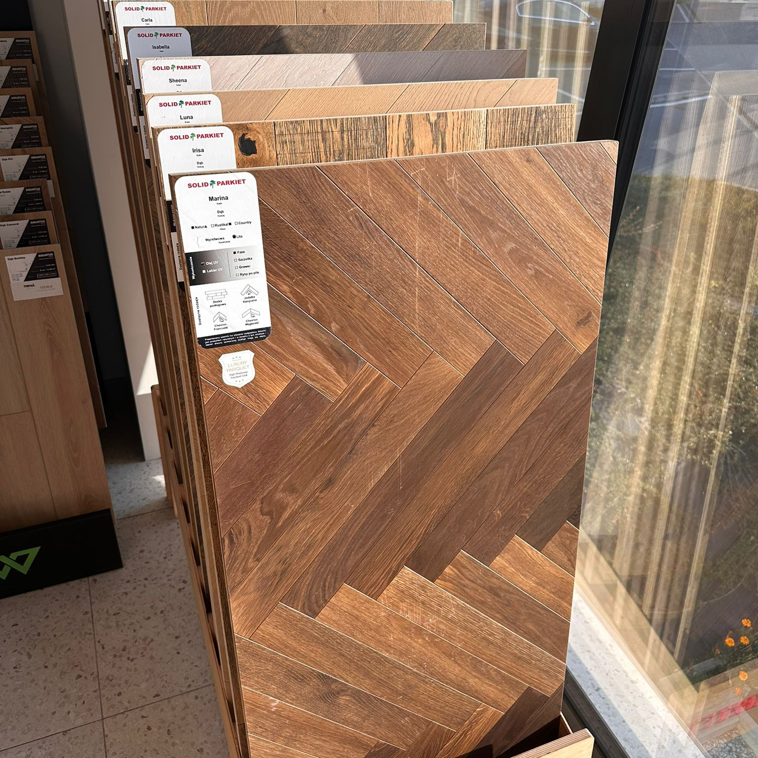 Ekspozycja próbek paneli podłogowych marki Solid Parkiet, różne odcienie brązu, wzór jodełki. Naturalne światło, widok z bliska na tekstury drewna.