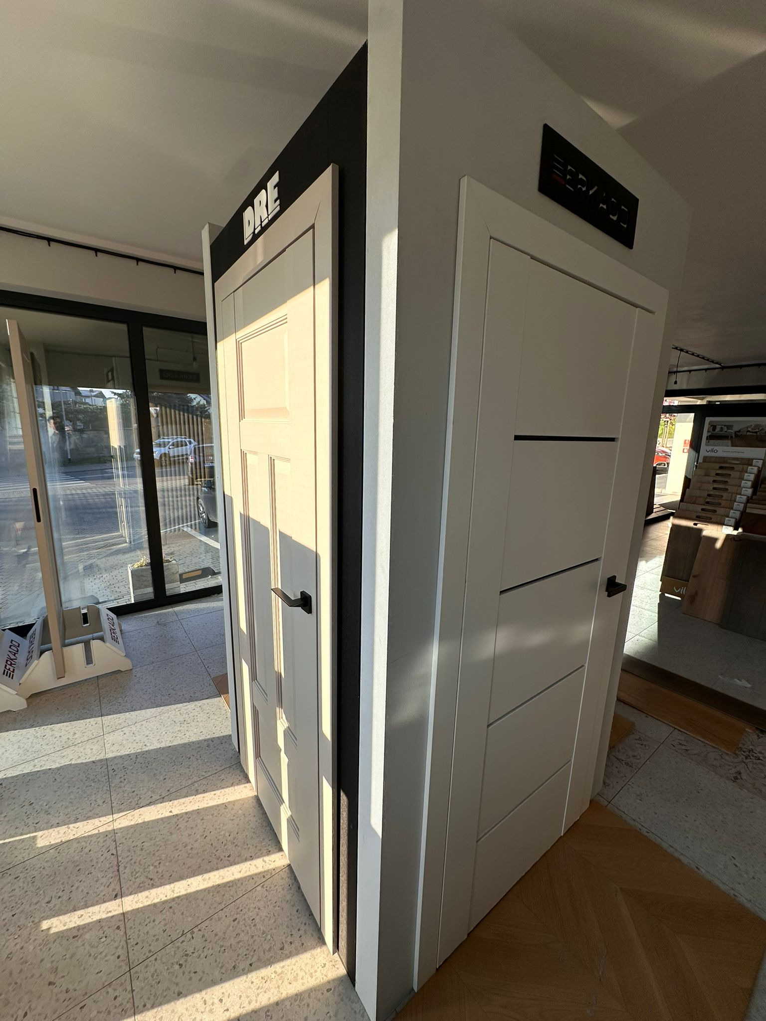 Ekspozycja dwóch modeli drzwi wewnętrznych: klasyczne DRE z frezowaniami i nowoczesne ERKADO z poziomymi intarsjami. W tle widoczna podłoga i okno.