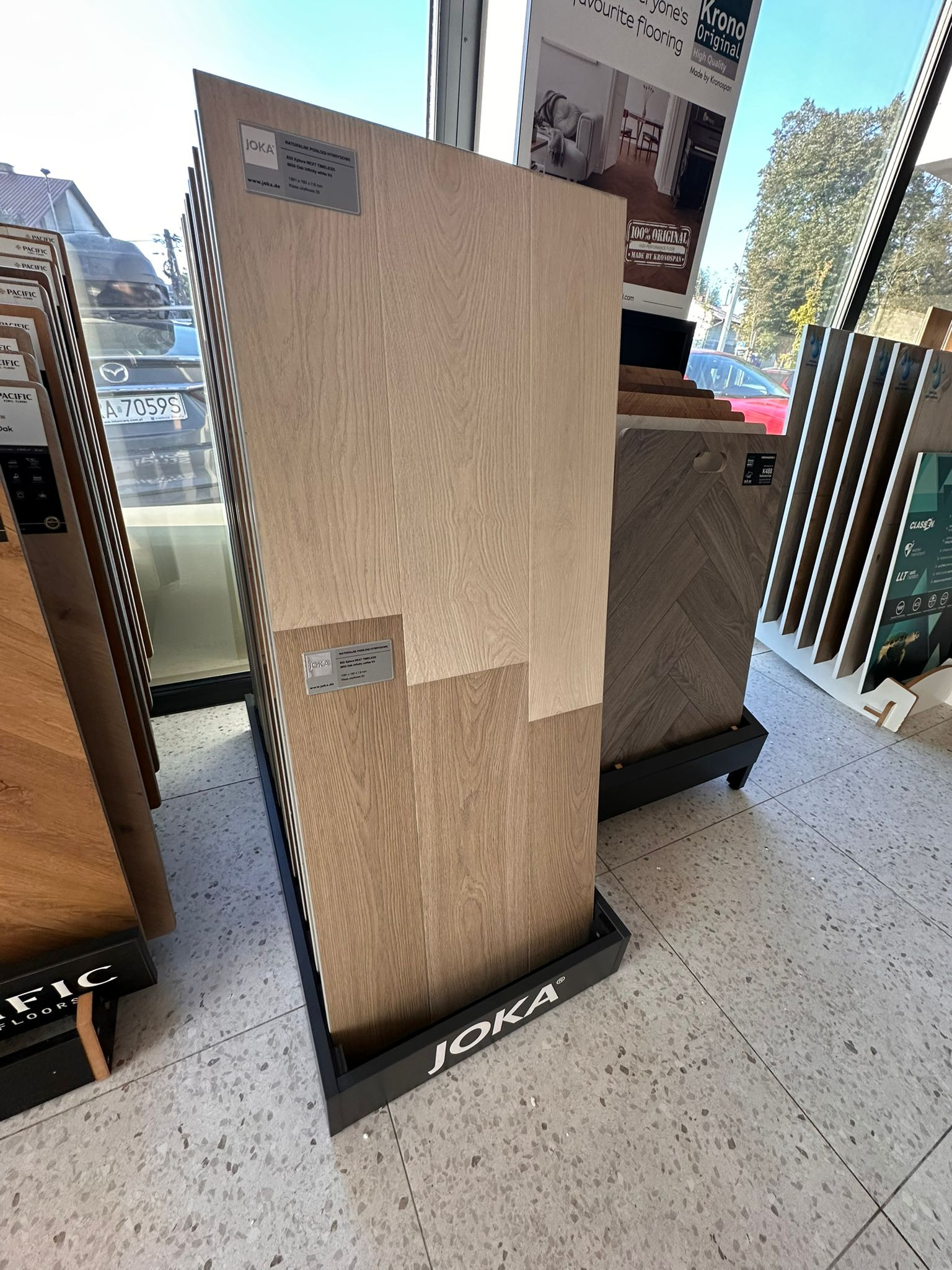 Ekspozycja paneli podłogowych Joka w salonie, prezentująca różne wzory i odcienie drewna. Widoczne próbki w stojakach, jasne oświetlenie, wzorzysta podłoga w tle.