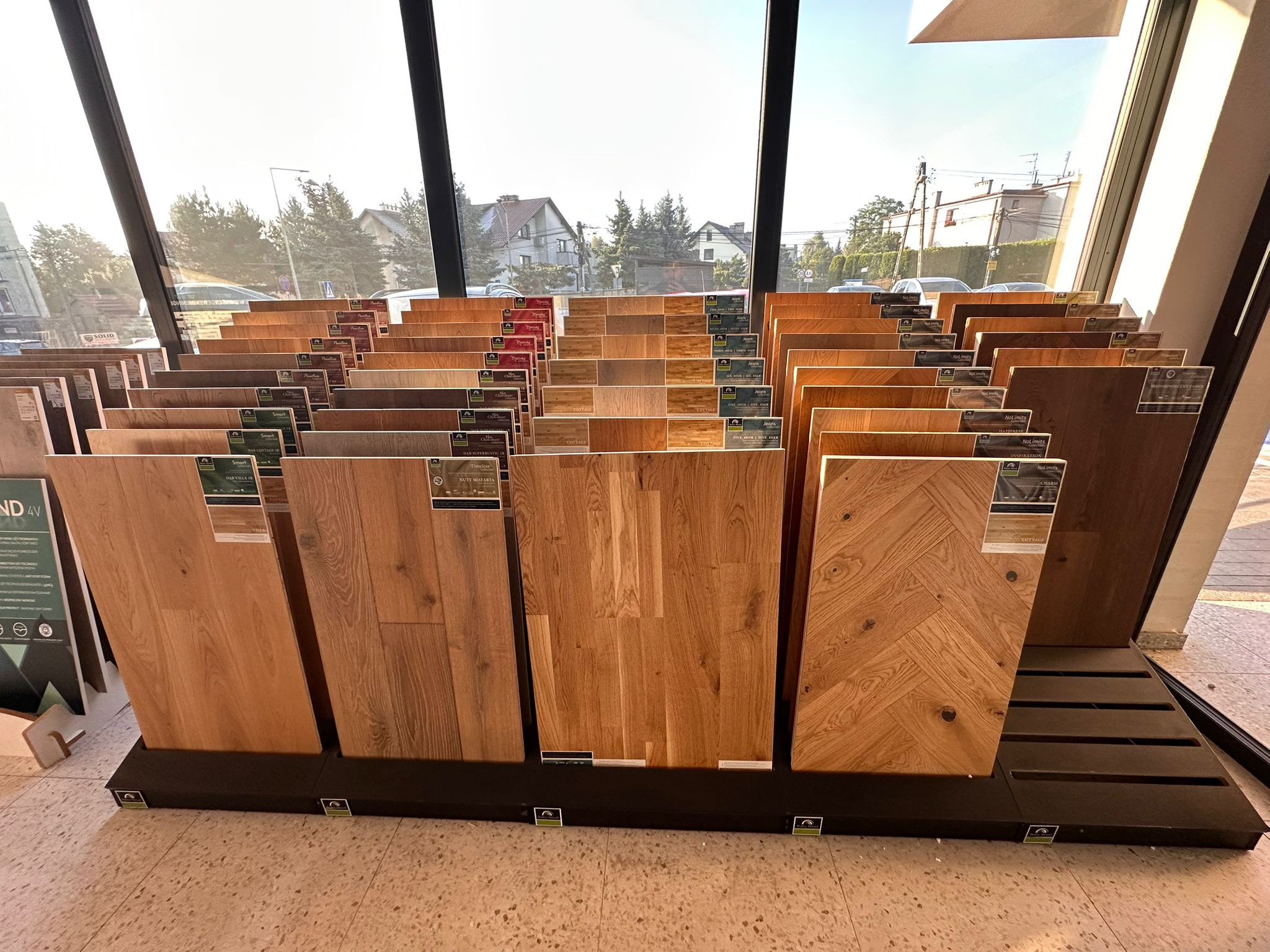 Ekspozycja różnorodnych paneli podłogowych w salonie, prezentująca bogatą gamę kolorów i wzorów drewna, z widokiem na ulicę przez okno w tle.