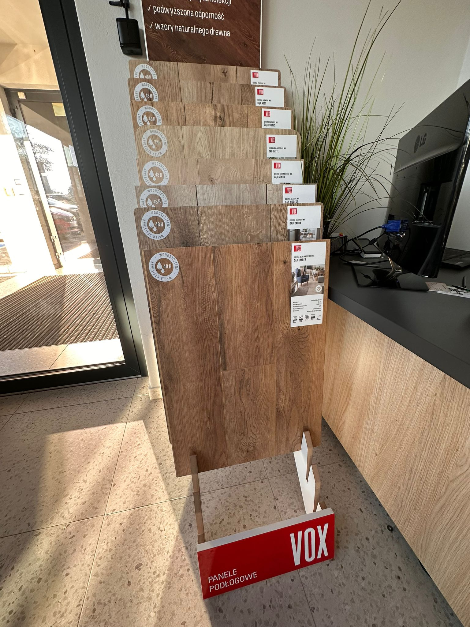 Ekspozycja paneli podłogowych Vox w sklepie: różne odcienie drewna na stojaku, etykiety z nazwami kolekcji i logo. Widoczny fragment podłogi i wejście do sklepu.