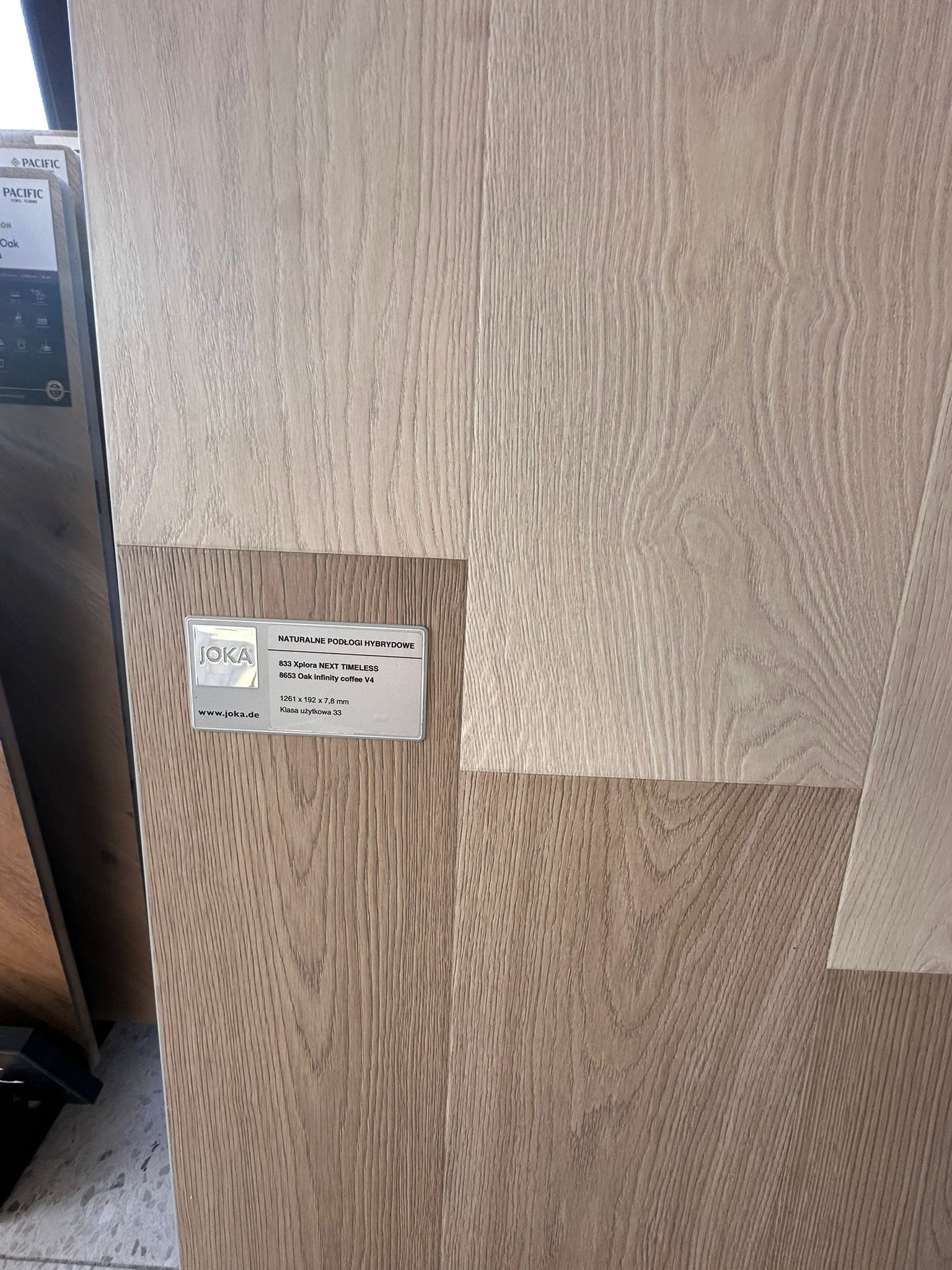 Próbki paneli podłogowych JOKA, w odcieniach jasnego drewna, z widoczną etykietą informacyjną o parametrach technicznych i nazwie modelu: Xplora NEXT TIMELESS.