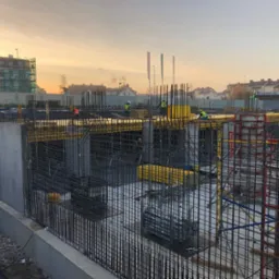 Zbrojenie fundamentów budynku wielorodzinnego w trakcie realizacji. Widoczne stalowe pręty i formy szalunkowe, pracownicy w odblaskowych kamizelkach w tle, ciepłe światło.