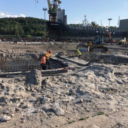 Stratos Budownictwo - Praca zbrojarska na placu budowy w porcie: zbrojenie fundamentów, pracownicy w kamizelkach, ciężki sprzęt i statek Wilson Astakos w tle, słoneczny dzień.