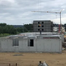 Surowy stan budynku żelbetowego z widocznym fundamentem i ścianami, na tle placu budowy z dźwigiem i budynkiem w rusztowaniach. Robotnik na dachu.