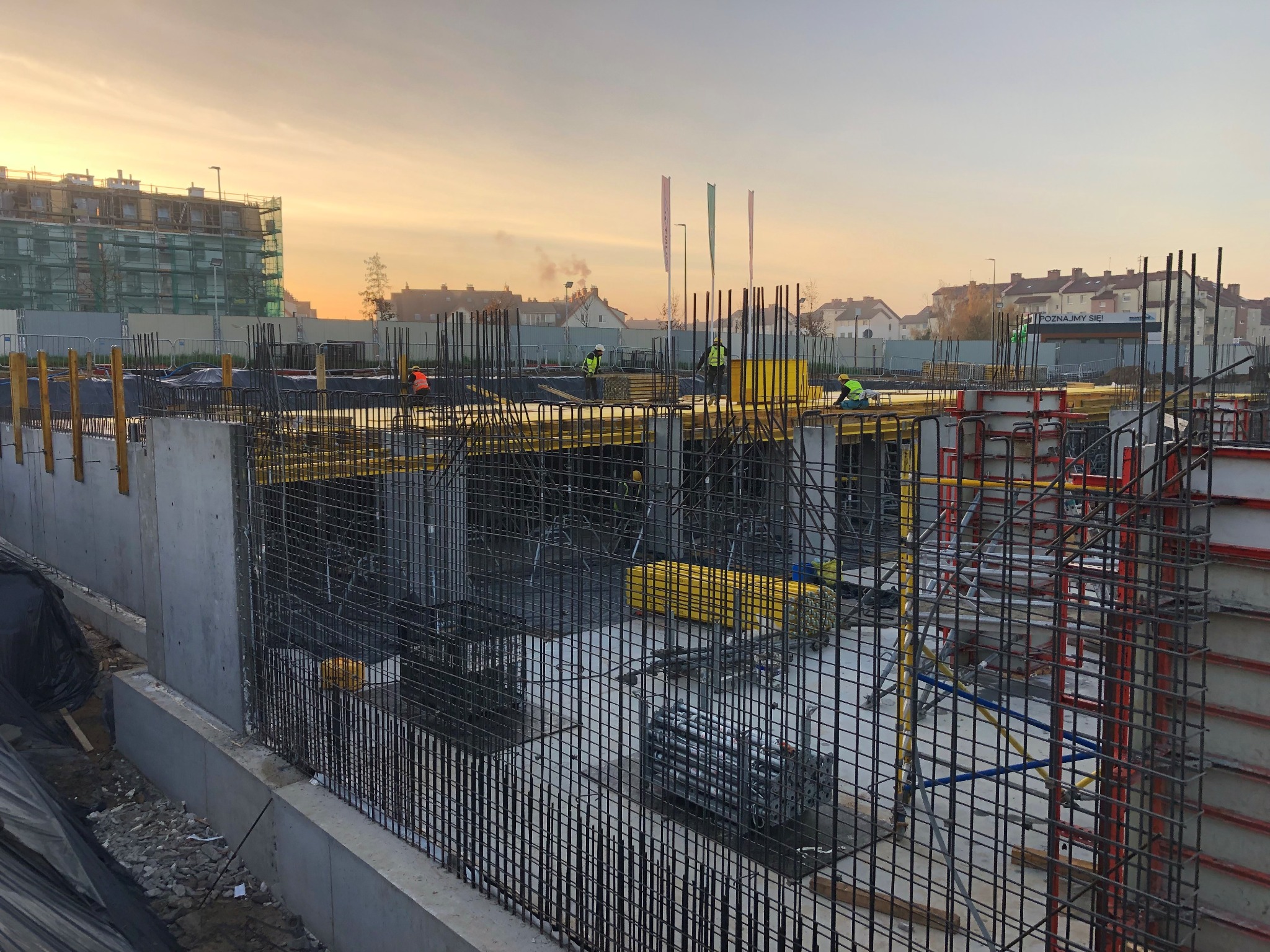 Zbrojenie fundamentów budynku wielorodzinnego w trakcie realizacji. Widoczne stalowe pręty i formy szalunkowe, pracownicy w odblaskowych kamizelkach w tle, ciepłe światło.