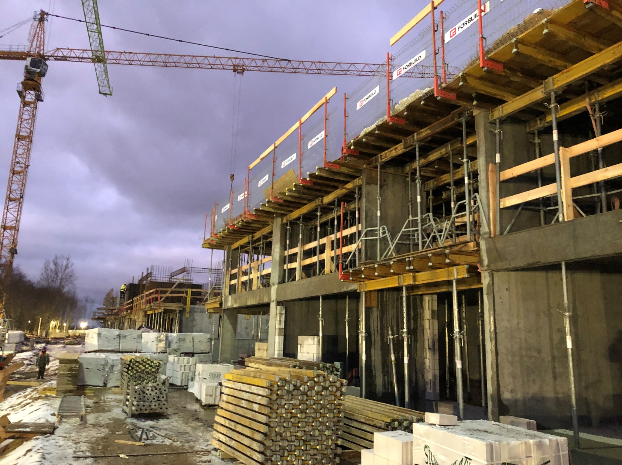 Wczesny etap budowy budynku żelbetowego z szalunkami stropowymi i widocznym żurawiem wieżowym na tle pochmurnego nieba. Składowane materiały budowlane.
